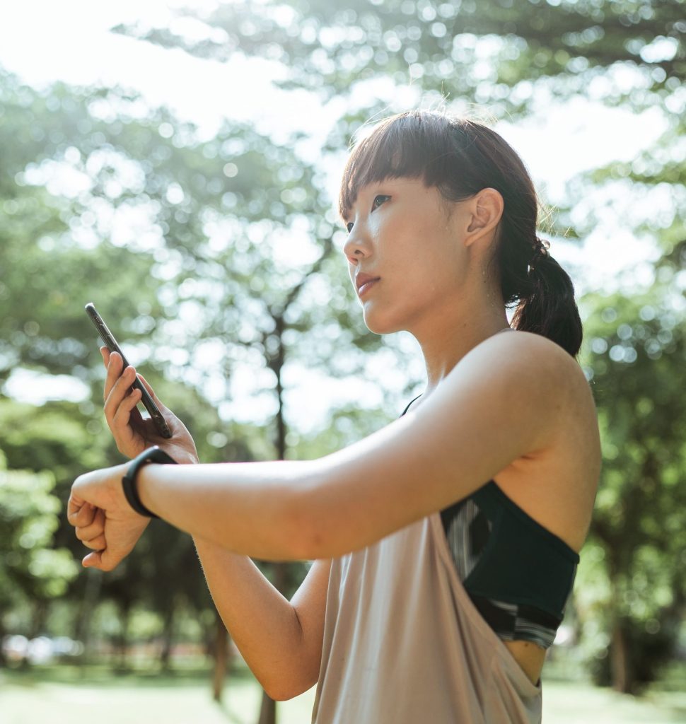 From below of Asian female wearing sportswear standing in park and using smart watch with mobile phone